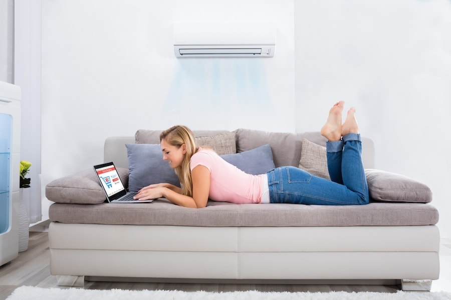 Young Happy Woman Lying Under Air Conditioner On Couch Using Laptop At Home.